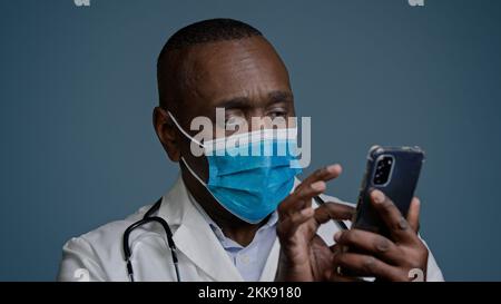 Close-up pensive thoughtful african american man doctor in protective mask holds phone studies new information on Internet ponders uses medical mobile Stock Photo