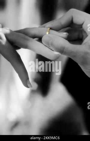 Black And White Photo Of A Groom Slipping A Gold Wedding Band On A Bride’s Finger During The Wedding Ceremony Representing The Promises Made During We Stock Photo