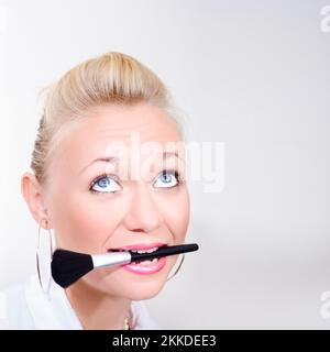 Comical Beauty Portrait On The Face Of A Makeup Woman With Bright Pink Lips And Wide Eyes Holding Make-Up Brush In Mouth Stock Photo