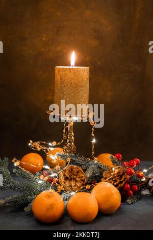 A golden candle burns on a candillabra on a black background. In the foreground are tangerines and spruce branches with cones. Light garlands. Stock Photo