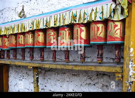 A row of colorful identical painted prayer wheels with gold lettering and carved wooden handles lined up along a white plaster wall in Thimphu, Bhutan Stock Photo