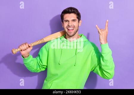 Portrait of funny crazy rock roll lover guy wear green hoodie excited hold baseball bat excited metal sign isolated on violet color background Stock Photo