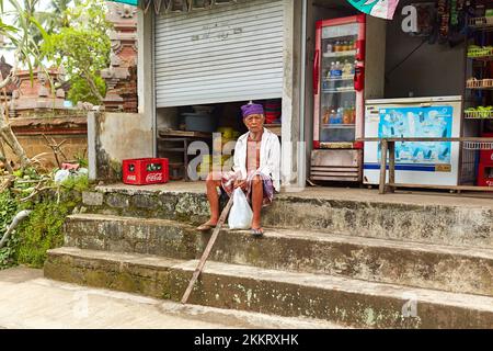 An elderly man in a sterno sits on the steps outside a grocery store. Bali, Indonesia - 03.22.2018 Stock Photo