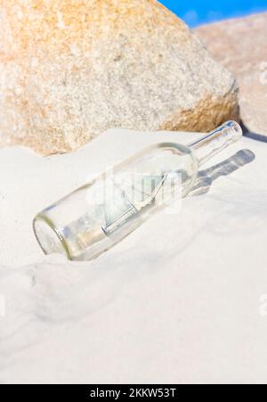 Glass Bottle Sits On In The Sand Of A Rocky Beach Shore Containing A Sailing Ship And Water In A Stranded Shipwreck Concept Stock Photo
