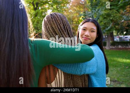 Three female friends embracing at a public park, while one of them turns to the camera smiling. Stock Photo