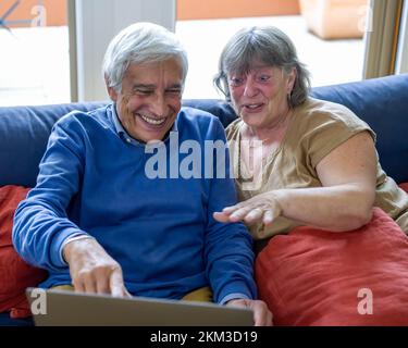 Elderly couple making a remote video call with grandchildren, retirees using a laptop and internet connections for shopping, grandparents watching som Stock Photo