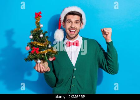 Photo of positive lucky man wear x-mas green cardigan rising fist holding pine isolated blue color background Stock Photo