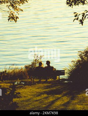 A vertical shot of a couple sitting on a bench against a lake at golden hour Stock Photo