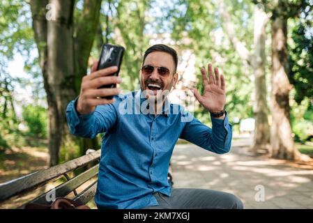 Delighted adult man, making a self portrait, waring sunglasses, sitting in the park, smiling for the picture. Stock Photo