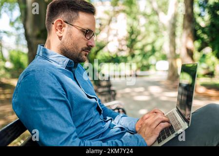 Focused adult man with eyeglasses, working hard and serious, using laptop outside, side shot. Stock Photo