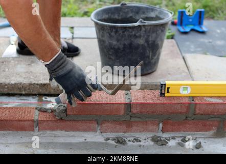 Building a wall of red bricks. Male hands holding brick trowel and level measurement tool. Masonry work in details. Construction site close up photo. Stock Photo