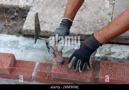 Building a wall of red bricks. Male hands holding brick trowel and level measurement tool. Masonry work in details. Construction site close up photo. Stock Photo
