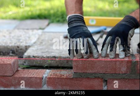 Building a wall of red bricks. Male hands holding brick trowel and level measurement tool. Masonry work in details. Construction site close up photo. Stock Photo