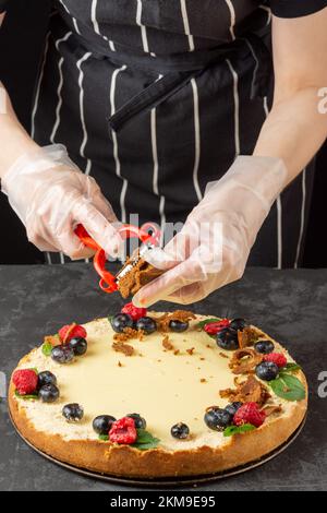 A female chef decorates a homemade classic cheesecake with fresh berries and grated salted cheese on a dark background Stock Photo