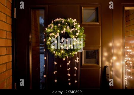Green color spruce tree tips Christmas Wreath with warm lights and white felt pom-poms on home entryway brown metal door with tiny windows outdoors. Stock Photo