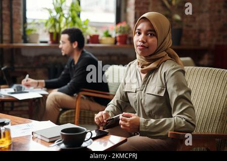 Young pretty businesswoman in hijab sitting by table in front of camera and looking at you while organizing work against colleague Stock Photo