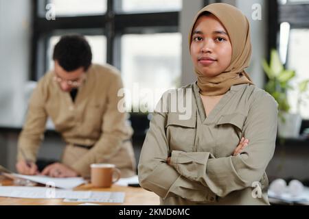 Young confident Muslim businesswoman in hijab with her arms crossed on ches looking at camera against male colleague signing papers Stock Photo