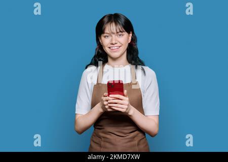 Smiling young female staff in apron using smartphone, on blue background Stock Photo
