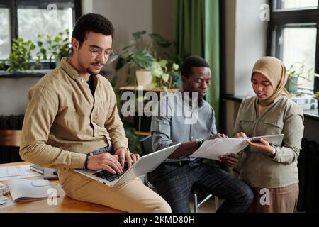 Young broker with laptop on his knees sitting on desk and networking against two intercultural colleagues discussing financial documents Stock Photo