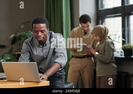 Young African American businessman bending over desk with laptop and networking against two intercultural colleagues using tablet Stock Photo