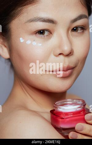 young woman with facial cream jar and eye cream on face against blue background. Stock Photo