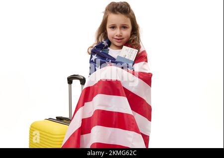 Lovely little girl wrapped in USA flag, holding air ticket and passport while standing with suitcase on white background Stock Photo