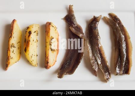Overhead view of a minimalist appetizer of three baked rosemary potatoes and three anchovies arranged in a row on a white plate ready to eat. Stock Photo