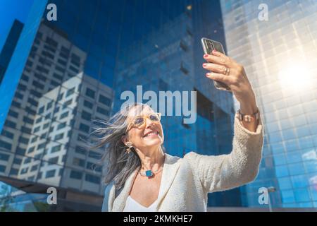 Happy businesswoman smiling and taking a selfie with mobile phone in front of business buildings. Stock Photo