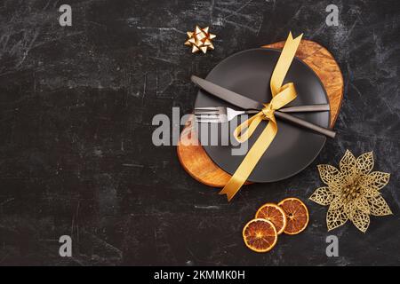 Black table setting with a plate on cutting board, fork and knife with golden bow, Christmas decorations and orange slices. Top view, flat lay, copy s Stock Photo