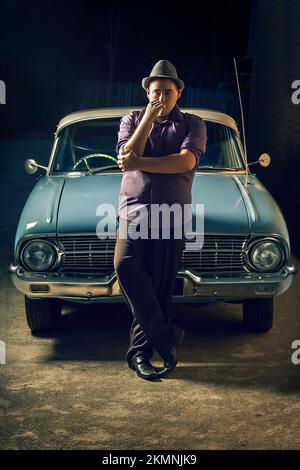 Pondering private eye investigator standing beside old classic car with smoke in hand awaiting a nightly rendezvous. Film noir concept Stock Photo
