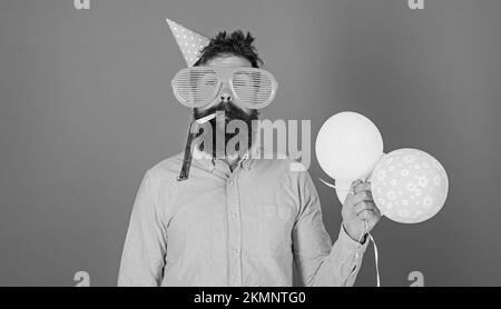 Celebration concept. Man with beard and mustache on calm face blows into party horn, blue background. Hipster in giant sunglasses celebrating birthday Stock Photo