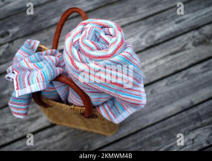 Heading out for a day at the beach - woven beach tote with leather straps; with colorful beach towels, one rolled, one loose, on rough, rustic, weathe Stock Photo