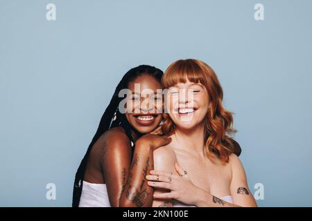 Women with different skin tones smiling happily while standing together. Two cheerful young women feeling comfortable in their own skin. Body positive Stock Photo