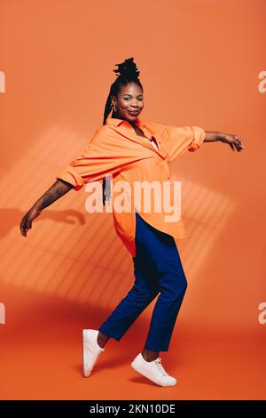 Fashionable black woman looking at the camera while dancing in casual clothing. Happy young woman having fun against an orange background. Stylish you Stock Photo