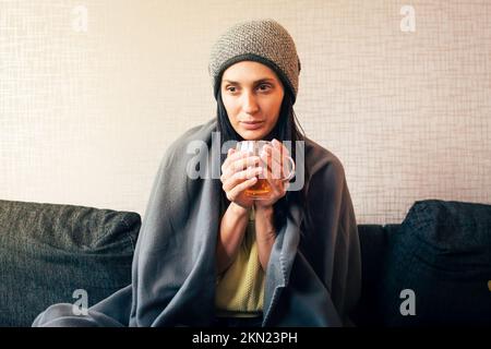 Caucasian Worried sad woman sits under blanket on sofa with cup of tea, Rising costs in private households for gas bill due to inflation and war, Ener Stock Photo