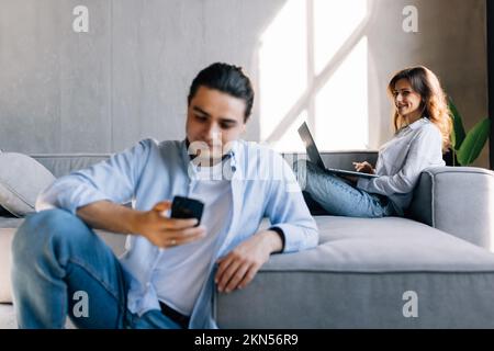 Joyful man and woman sitting together on couch in gray interior while using laptop and smartphone Stock Photo