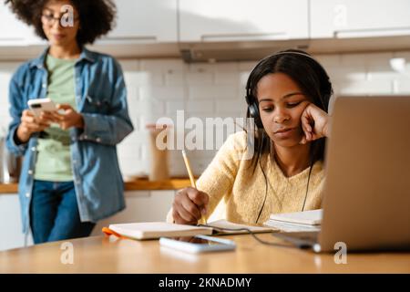Black girl writing in exercise book while doing homework with laptop at home Stock Photo