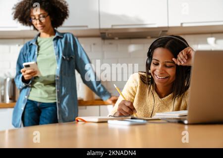 Black girl writing in exercise book while doing homework with laptop at home Stock Photo