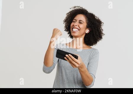 Young hispanic woman in earphones playing online game on cellphone isolated over blue background Stock Photo