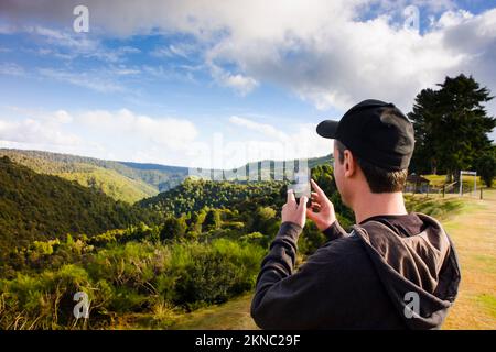 Horizontal photo of traveller man taking scenic picture with mobile smartphone of the mountains of Tarkine reserve from mount Waratah, North Tasmania, Stock Photo