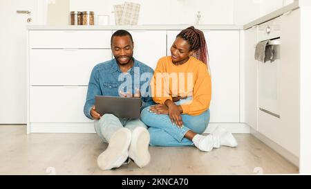 Excited african american spouses with laptop sitting on floor in kitchen, shopping online or browsing internet Stock Photo
