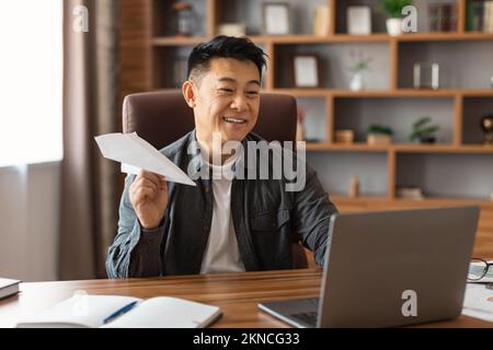 Smiling middle aged asian male making airplane with paper, planning strategy at table with laptop Stock Photo