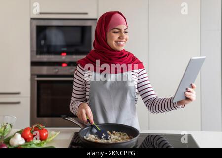 Happy Muslim Woman In Hijab Using Digital Tablet In Kitchen Stock Photo