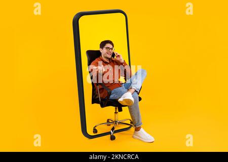Cheerful millennial european guy in glasses sits on armchair call by phone talk in big smartphone Stock Photo