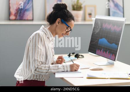 Happy businesswoman taking notes and analyzing indexes, financial charts trading online, graph on pc screen Stock Photo