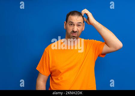 Miserable and gloomy guy with messy hair, frowning and looking unsatisfied while scratching head, standing over blue background. Something heavy fell Stock Photo