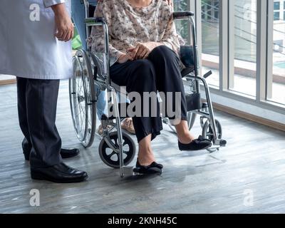 Senior woman patient person sits in wheelchair on the floor while man doctor in white coat standing beside to visit her inside the waiting room in hos Stock Photo