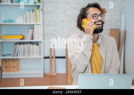 Young creative entrepreneur talking on the phone laughing smiling in his modern workspace at home. Stock Photo