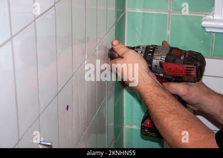 Dirty hands of plumber using a drill to create new holes in green and white tile in old fashioned bathroom Stock Photo
