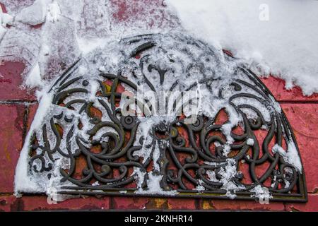 Ornate rubber welcome door mat in snow with ice and shoe prints on grungy painted red brick outdoor surface - top down view - closeup Stock Photo